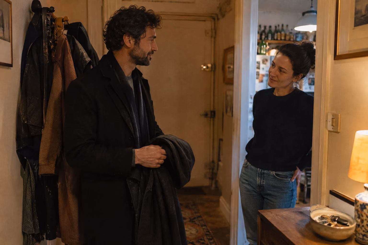 Dans l’entrée d’un appartement le soir, une personne rentre son manteau à la main tandis que son partenaire l’accueille d’un sourire pensif